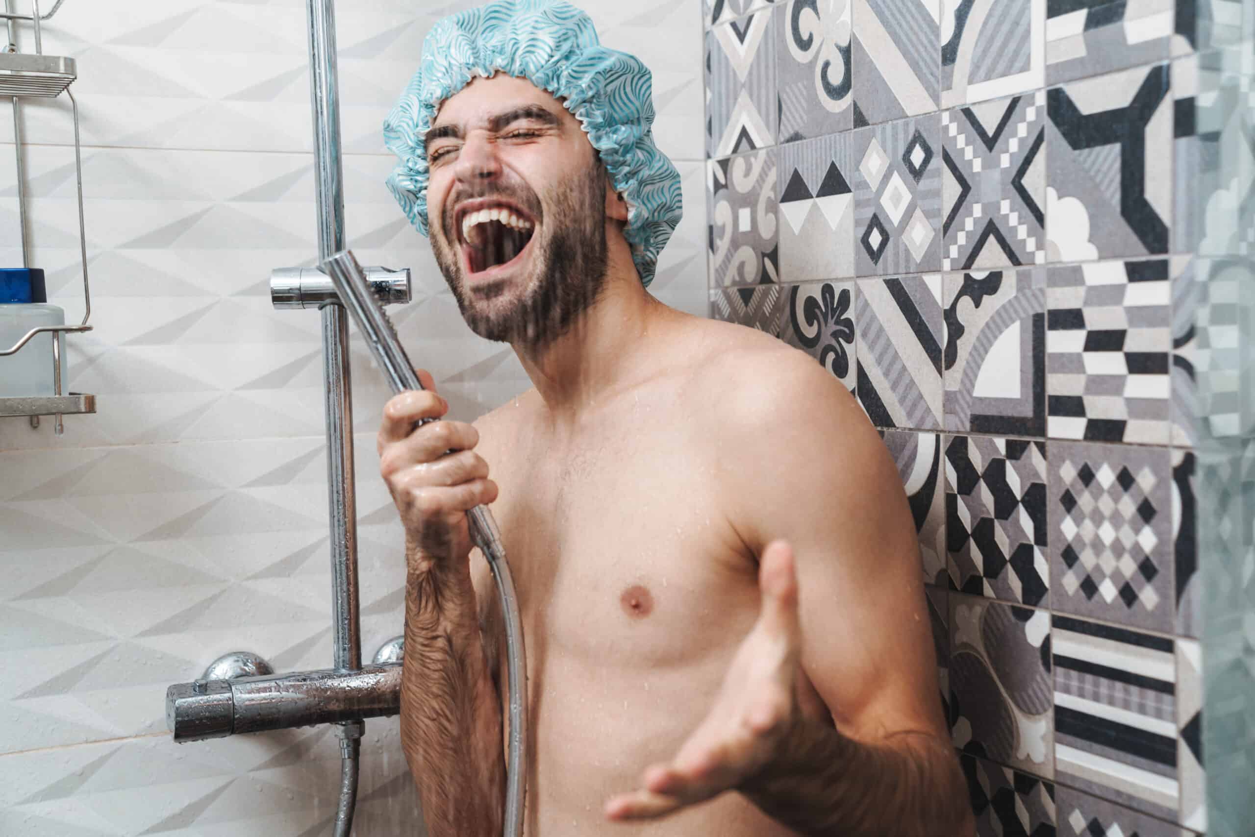 Attractive young cheerful man singing while washing in the shower, wearing shower cap and holding shower head